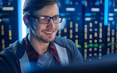 Experte für Systemhärtung | Mann mit Brille sitzt vor einem Computer, lacht und im Hintergrund sind Lichter von Servern zu sehen