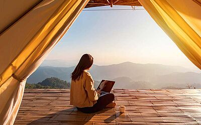 Workation | Blick aus einem Zelt auf eine Frau arbeitet am Laptop, im Hintergrund Berge, Wald und Sonne